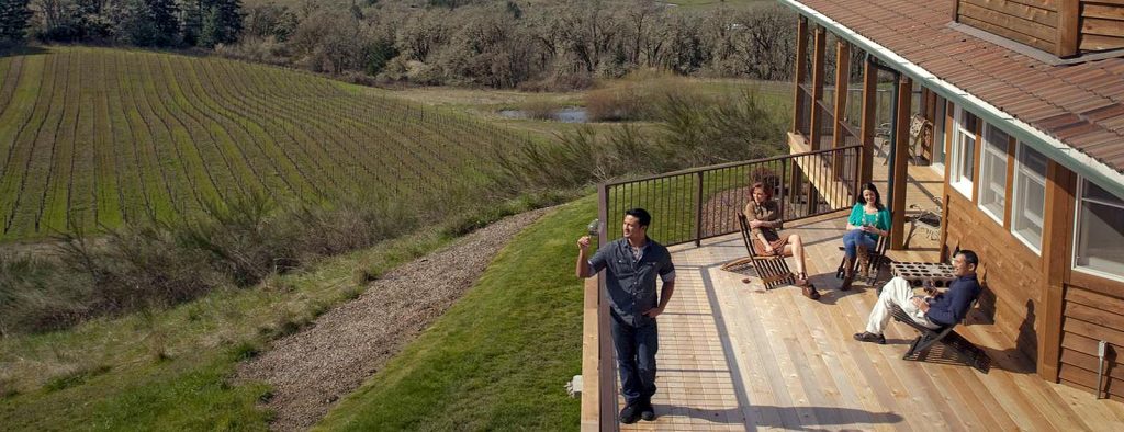 Three people sit on a wooden deck overlooking a hillside vineyard and landscape on a sunny day.