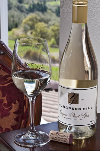 A bottle of Youngberg Hill Oregon Pinot Gris, a glass of white wine, and a cork sit on a table by a window.