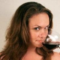 A woman with long brown hair looks toward the camera while holding a glass of red wine near her mouth.