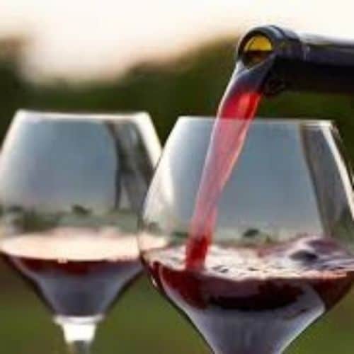 Red wine is being poured from a bottle into a glass, with another glass of red wine nearby against a blurred backdrop.