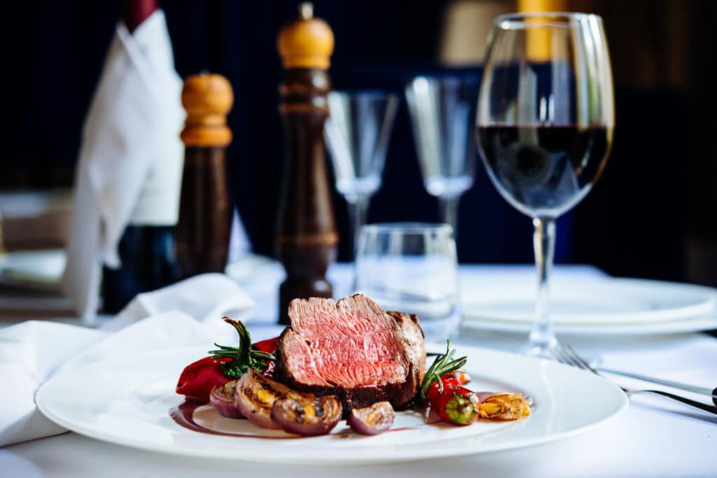 A plated steak with vegetables sits on a table set with wine, pepper grinders, and empty glasses.
