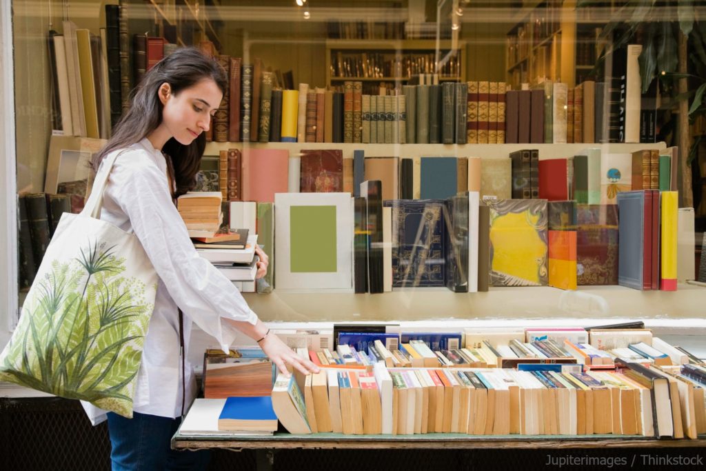A woman with a tote bag browses a table of books in front of a bookstore window filled with shelves of books.