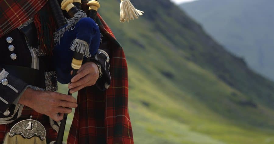 A close-up of a person in traditional Scottish attire playing bagpipes, with a hillside in the background.