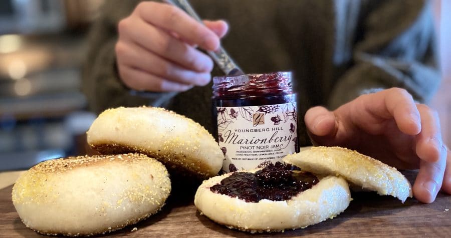 Homemade Jam Featuring our Oregon Wines 1 A person spreads marionberry jam on an English muffin on a wooden board with other muffin halves.