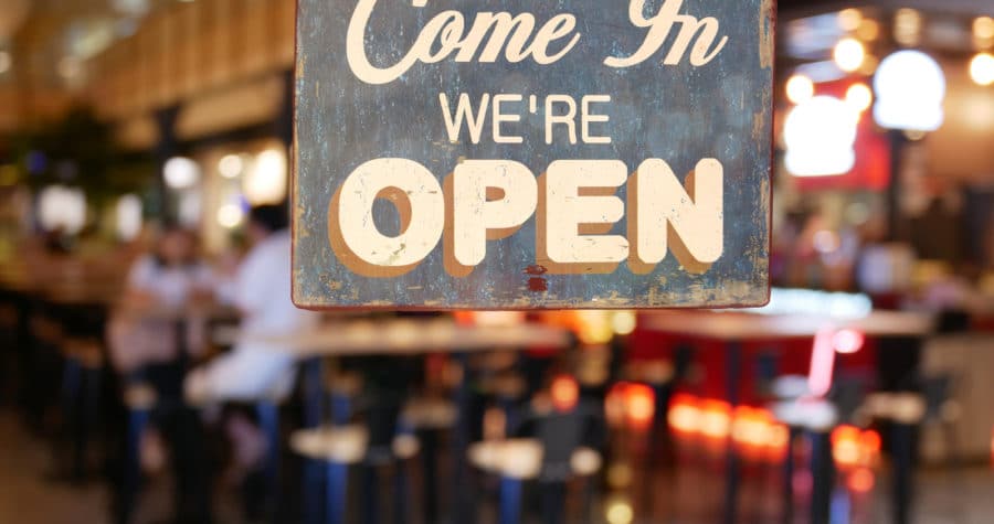 A sign reading "Come In WE'RE OPEN" hangs in the foreground of a blurred restaurant or bar.