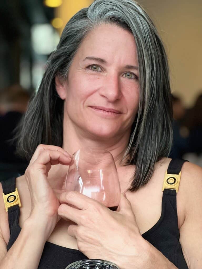 A woman with shoulder-length gray hair holds a wine glass, wearing a black tank top with gold hardware on the straps.